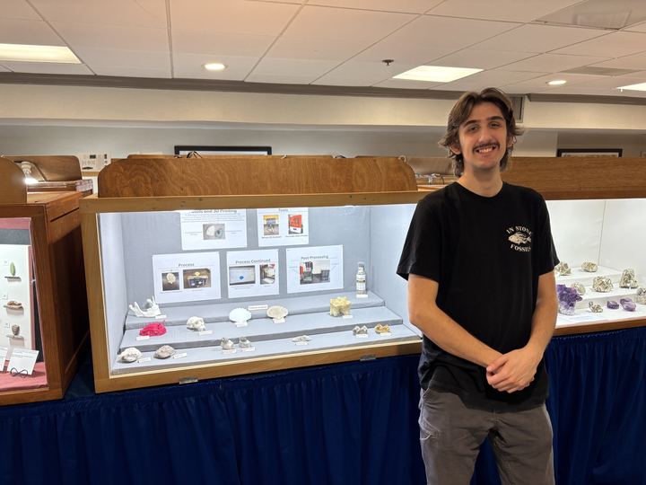 Luke standing beside a museum display case he assembled, with panels on fossils and 3D printing, tools, process and post-processing, and printed specimens laid out on risers