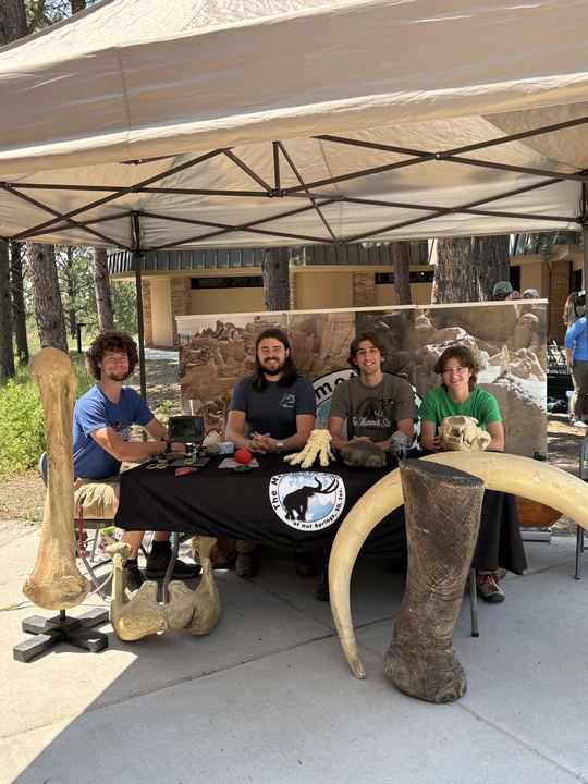Four interns behind an outdoor handling table at Jewel Cave National Monument with a mammoth femur, pelvis, tusks and a skull cast set out for visitors