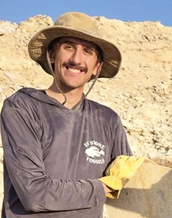 Luke in a sun hat and quarry shirt holding work gloves, a quarry wall behind him